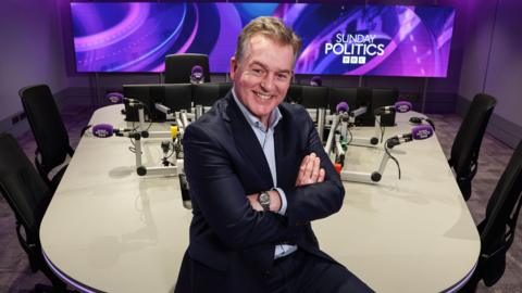 Mark Carruthers is sitting against a desk in a studio. His arms are folded. He's wearing a navy suit with alight blue shirt and a silver watch. Purple mics at each desk state Sunday Politics and a graphic on a screen int he background states Sunday Politics BBC. 