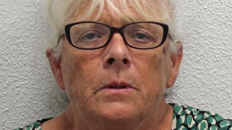 Deborah Mason, a woman with white hair and wearing dark rimmed glasses. She is standing against a white background and wearing a green, white and black patterned top