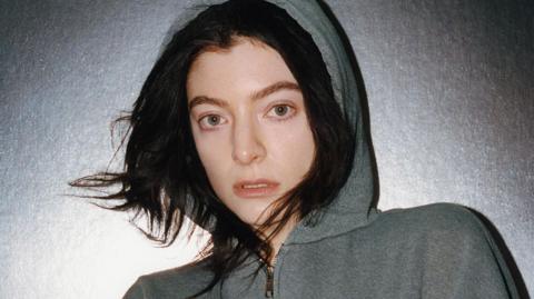Lorde. She is pictured against a silver background wearing a grey hoodie with the hood up. She has long dark hair which is peeking out from underneath the hood.
