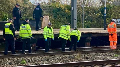 A group of transport police officers walk along the track, looking closely for clues. They wear high-vis jackets over black hoodies and black trousers., One railway worker dressed in orange walks ahead of them.