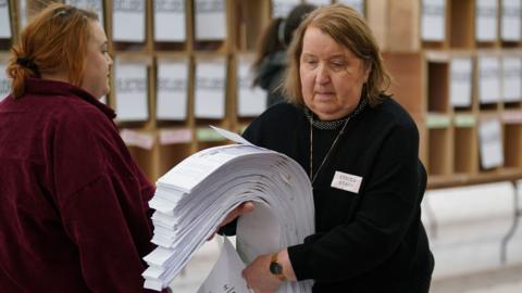 Woman carrying a stack of ballot papers. She has brown hair and is wearing a black jumper. She has a badge on which says count staff