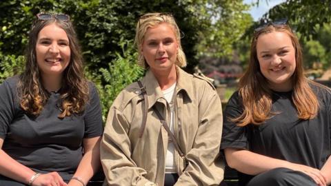 All three women in their late twenties and early thirties are sat on a bench in a park. Cari is on the left wearing a black outfit. Mererid is in the middle wearing a light beige coat and Nia is on the right in a black outfit.
