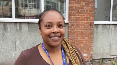 Joanne Alexander looks straight ahead, towards the camera, and is smiling. Her hair is tied back and she is wearing a brown top with a blue lanyard that has "staff" written on it in white capital letters. She is pictured with the exterior of a building behind her.