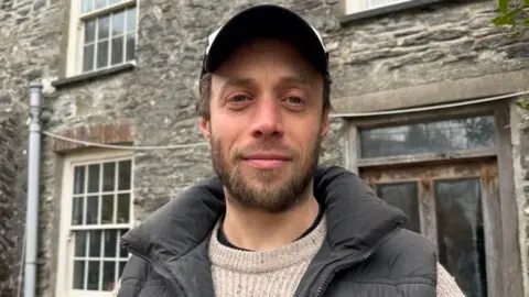Owen Shires yn sefyll o flaen ei gartref, sydd yn dy cerrig gyda drws pren