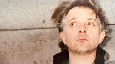 Comedian and actor Rik Mayall, standing with his back against a concrete wall. His hair, mid-brown with grey patches on the sides, is unkempt and he has a five o'clock shadow. He is looking up and behind the camera.