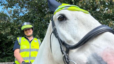 A woman wearing a high vis waist coat with a black helmet on that has a high vis strip around it, she's wearing a head cam pointing towards the camera and is sat on a white horse holding the reins. The horse has a sparkly head band around its ears and a small high vis hat on with his left eye looking towards the camera. 