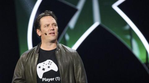 A close up of Phil Spencer at a conference with the Xbox logo in the background, he is wearing a black t shirt with a white Xbox controller on it and the white words Player (1) on it.