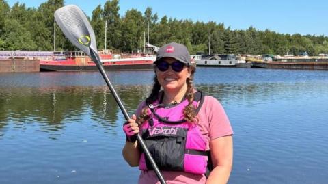 A woman in a pink T shirt and life vest, and a grey hat, stands on a paddle board holding a paddle in a dock
