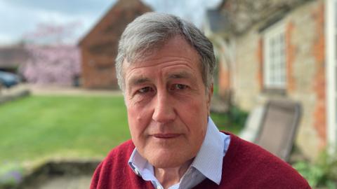 A man with grey hair wearing a blue striped shirt and red jumper looking directly at the camera. He is sitting in a garden in the background are chairs, grass and buildings.