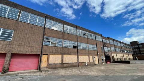 A large brick building with long rows of windows. The wide concrete yard in front is empty. The image has been taken on a sunny day with a blue sky overhead.