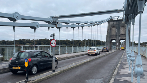 Three cars cross Menai Bridge with its huge blue grey metal hanger clearly visible, holding the road in place