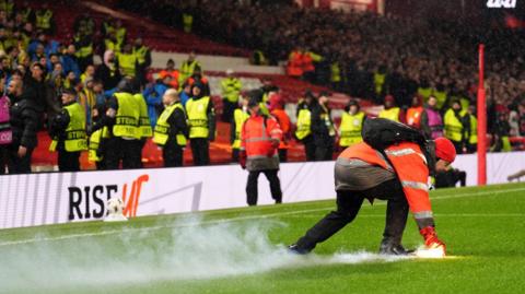 A man in an orange jacket picks up a lit flare in a red steel cage. He is stood on the side of a football pitch with fans and stewards behind him.
