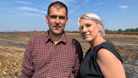 A man wearing a chequered shirt and woman wearing a dark top stand next to each other looking into the camera. They are in a field - with blackened and burnt crops behind them. It is a sunny day with blue sky.