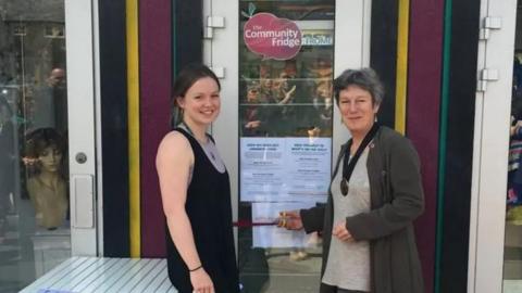 Esther Deeks and Ali Barclay open the first community fridge in Frome in 2016. They are standing next to the fridge and Ali is cutting the ribbon with scissors.