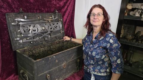 Auctioneer Ann Thorp stands next to a 17th Century iron-played strongbox, which has been opened to show details inside the item. Ann is wearing a blue floral shirt and has her right arm resting on the side of the box.
