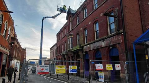 A tall red brick building can be seen but in front are metal fences with warning signs all over reading 'danger'. A crane can be seen as workers in hi-vis jackets.
