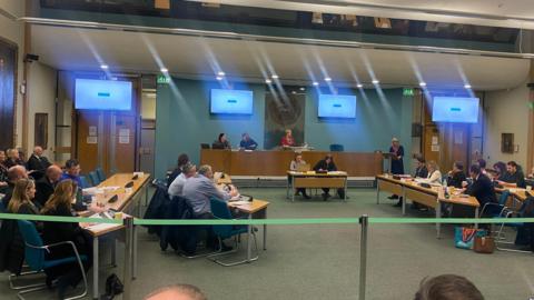 Hartlepool councillors sitting in rows at desks in a meeting chamber. There is a long desk on a podium at the top of the room, where the mayor sits. There is a lectern on the right side of the desk. Four TV screens are suspended from the ceiling.