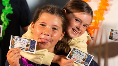 A close-up image of two girls holding 'Moana Exclusive Screening' cinema tickets. One is poking her tongue out. They are both smiling and looking into the camera.