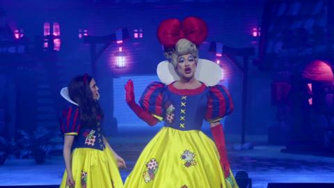 Jamie Alexander Wilson is the dame on the right, wearing a full yellow skirt, an attached blue and red shiny blouse, red gloves, a big blonde wig in an up do, a huge red bow, a white collar and exaggerated make-up with blue eye shadow and red lips. He holds his hand up in the face of Snow White on the left, who wears a similar outfit but without the make-up or bow. 