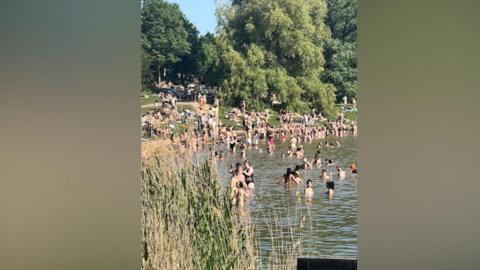 The image shows groups of swimmers in a non-bathing pool, with people gathered by the edge and people sitting on the grassy banks of the pool on Hampstead Heath.