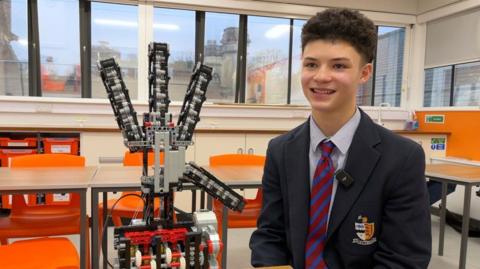 Jared wearing a school blazer, sitting next to his Lego hand and smiling at someone off camera. He is in a classroom with school tables and chairs.