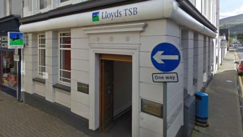 A closeup of the entrance and signage of the Ramsey Lloyds Bank Branch. It is a white building with a wooden doorframe and has the bank's name and logo above the door, on brass plaques either side, and on a stand alone sign to the left. There is a One-Way road sign on the pavement in front of it.