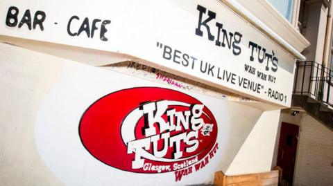 The entrance to a music venue. The club logo - KING TUT'S WAH WAH HUT is large, on white letters with a red background. The venue door is just next to the sign.