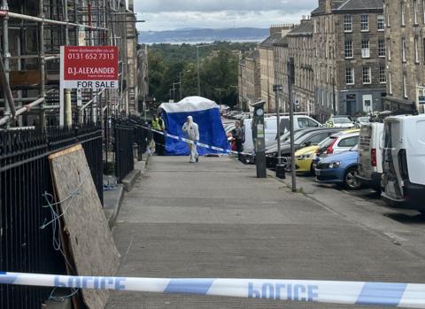 Street showing police tape, a blue police tent and scaffolding against a building