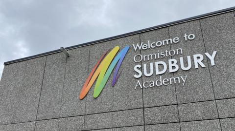 A general view of the exterior of Ormiston Sudbury Academy. The grey coloured building has the name of the school written on it white writing that reads 'Welcome to Ormiston Sudbury Academy'.