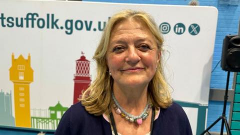 A woman is standing in front of a county council poster and speaker. She is smiling at the camera and is wearing a blue cardigan and a green and pink necklace. 