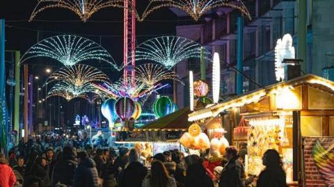 A Christmas market with lots of people lining a street with brightly lit stalls on the right side and multi-coloured lights lining the street
