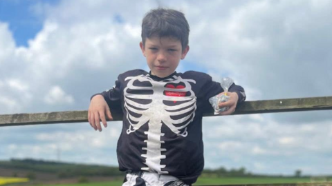 Travis is leaning against a wooden fence. He has short brown hair and is wearing what black top and trousers with a skeleton design on them. He is holding a small plastic bag in his left hand.