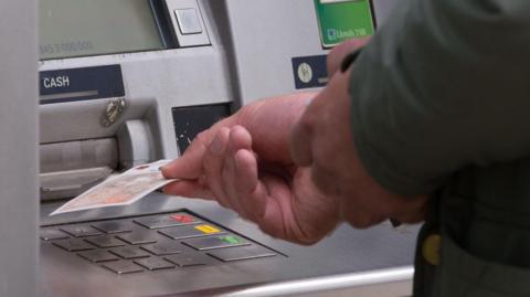 A person's hand with a green long sleeve top withdrawing a £10 note from an ATM machine