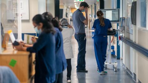 Doctors and nurses walking and standing in a busy hospital corridor.