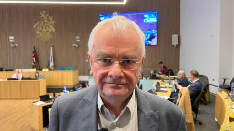 Jeremy Hilton looks directly at the camera at close quarters as he stands in a modern council chambers. He is an older man with grey hair and thin-rimmed, rectangular glasses. He is wearing a grey blazer and with a pale shirt beneath it. He has a half-smile on his face. Other council members can be seen sitting behind him and there is a large screen in the room.