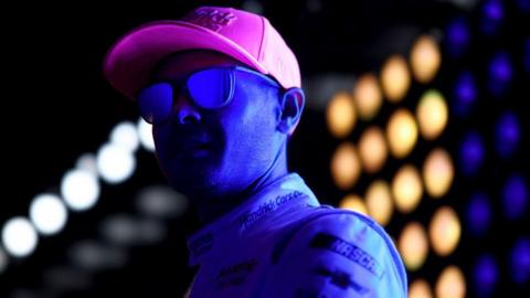 Kyle Larson waits backstage during pre-race ceremonies for the NASCAR Cup Series in Concord, North Carolina. He wears a bright pink cap and a light-coloured racing suit with sponsor logos, under vivid blue lighting and a backdrop of glowing circular lights.