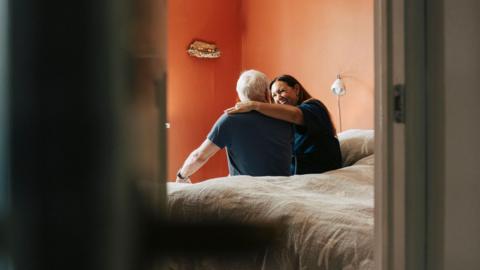 An elderly man sits on a bed with a female carer sitting beside him. She has 