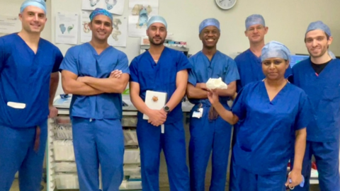 Seven people are wearing blue healthcare uniforms and looking at the camera. There are six men, near a wall that includes a clock, and a woman is in front of them.
