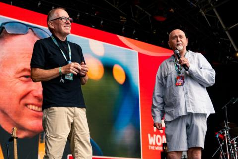 Two men stood smiling on a stage with lanyards on which say 'Womad'