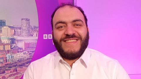 Nour Al-Baarini sat against a purple BBC backdrop, with a view of Birmingham's skyline. He is wearing a white shirt and smiling. 