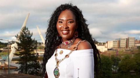 Shows Lilian Seenoi Barr in a white dress wearing the mayoral chain and a colourful necklace and standing against the backdrop of the peace bridge.
