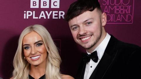 Project manager Jake Brown and former soldier Leanne Quigley in formal attire smiling at a photocall at the BBC. Jake is wearing a black bow tie.