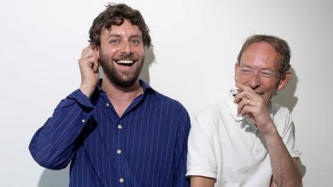 Felix White on the left earing a blue shirt. He has short wavy hair and a beard and is laughing. Steve Lamacq on the rightis wearing a white polo shirt and laughing.