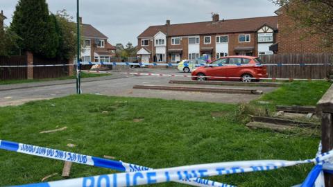 Richmond Court in Grangetown: a small cul-de-sac consisting of a cluster of red-brick houses. Police tape has been hung across the road to prevent access. A police car is parked outside a house which has a broken window.