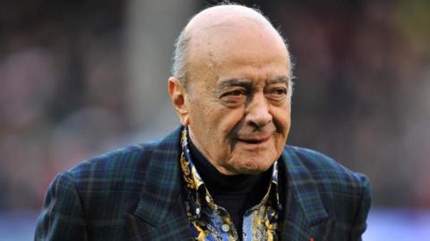 Mohamed Al Fayed wearing a dark green tartan jacket and black, blue and yellow printed shirt with a black turtle neck jumper underneath