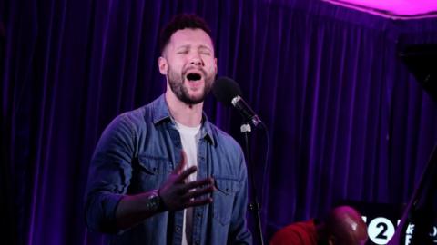 Calum Scott singing into a mic for Radio 2 with his hand in front of him wearing an unbuttoned denim shirt and white top with a purple curtain and pink lighting in the background