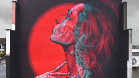 A 3D image of a woman with red and grey hair behind a red sun on the side of a building