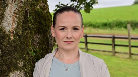 Anna Owens has black hair swept back from her forehead. She is standing beside a tree with fields in the background.