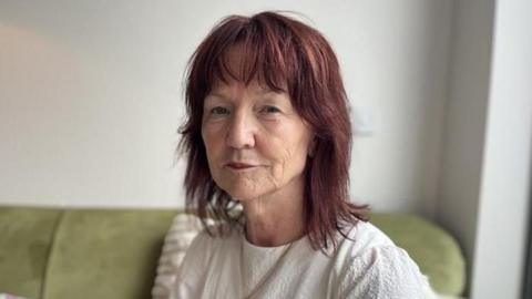 Afryl, who has a white top, looks at the camera. She has shoulder length red hair and is sitting on a green sofa. It is a head and shoulders shot.
