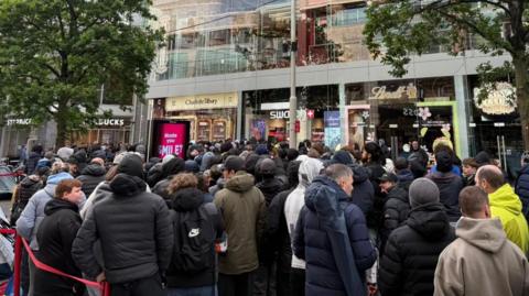 Hundreds of people wait in a queue snaking back from the shop entrance on a pedestrianised street. Glass fronted stores line the road on both sides.